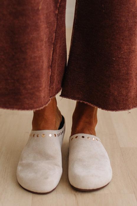 Close-up of a person wearing off-white suede mule slippers with metallic stud detailing, paired with rust-brown wide-leg pants on a light wood floor.