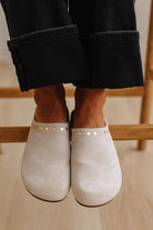 A close-up of a person wearing off-white suede mule shoes with gold-tone studs, paired with dark cuffed jeans, sitting on a wooden chair.