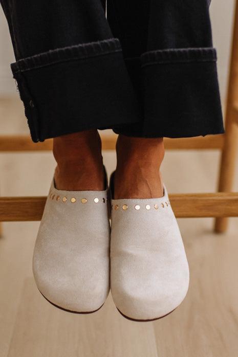 A close-up of a person wearing off-white suede mule shoes with gold-tone studs, paired with dark cuffed jeans, sitting on a wooden chair.