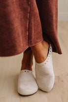 A close-up of a person wearing off-white slip-on mule shoes with gold-tone stud detailing around the collar, paired with a rust-brown maxi skirt on a light wood floor.