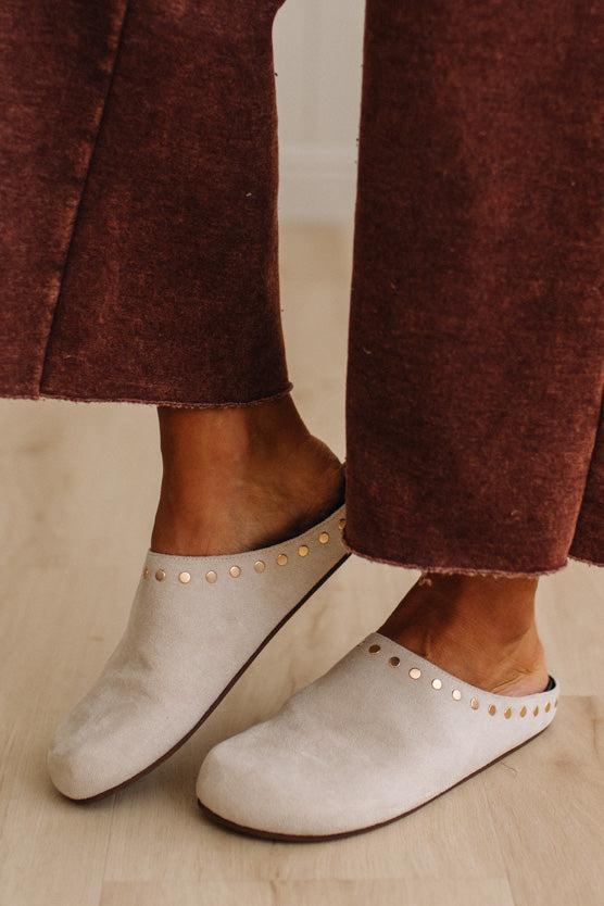 A close-up of a person wearing off-white suede mule slip-on shoes with gold-tone studs, paired with rust-brown wide-leg pants, standing on a light wood floor.