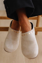 A close-up of a person wearing off-white suede clogs with gold-tone stud detailing around the opening, shown against a light wood floor and chair.