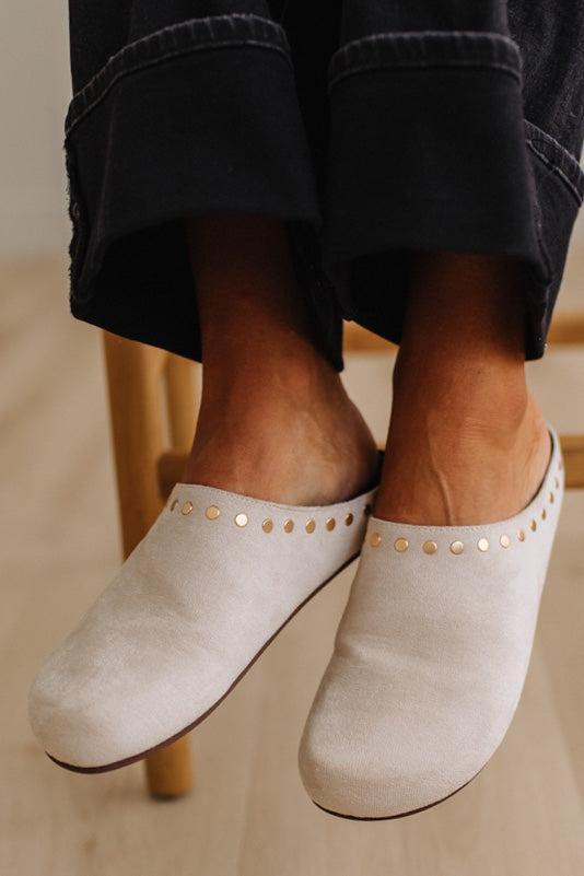 A close-up of a person wearing off-white suede mule slip-on shoes with gold-tone stud detailing along the top, paired with dark cuffed jeans.