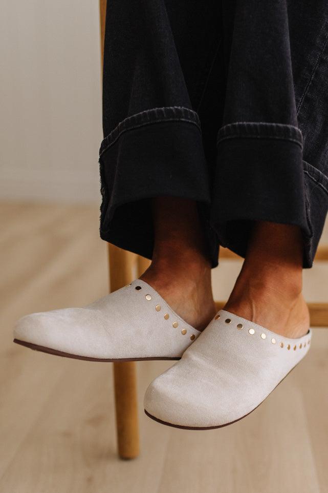 A close-up of a person wearing off-white suede mule slip-on shoes with gold-tone stud detailing along the side, paired with dark cuffed jeans, sitting on a wooden chair.