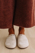 A close-up of a person wearing off-white suede clogs with small metal eyelets, standing on a light wood floor with rust-brown wide-leg pants visible above the feet.