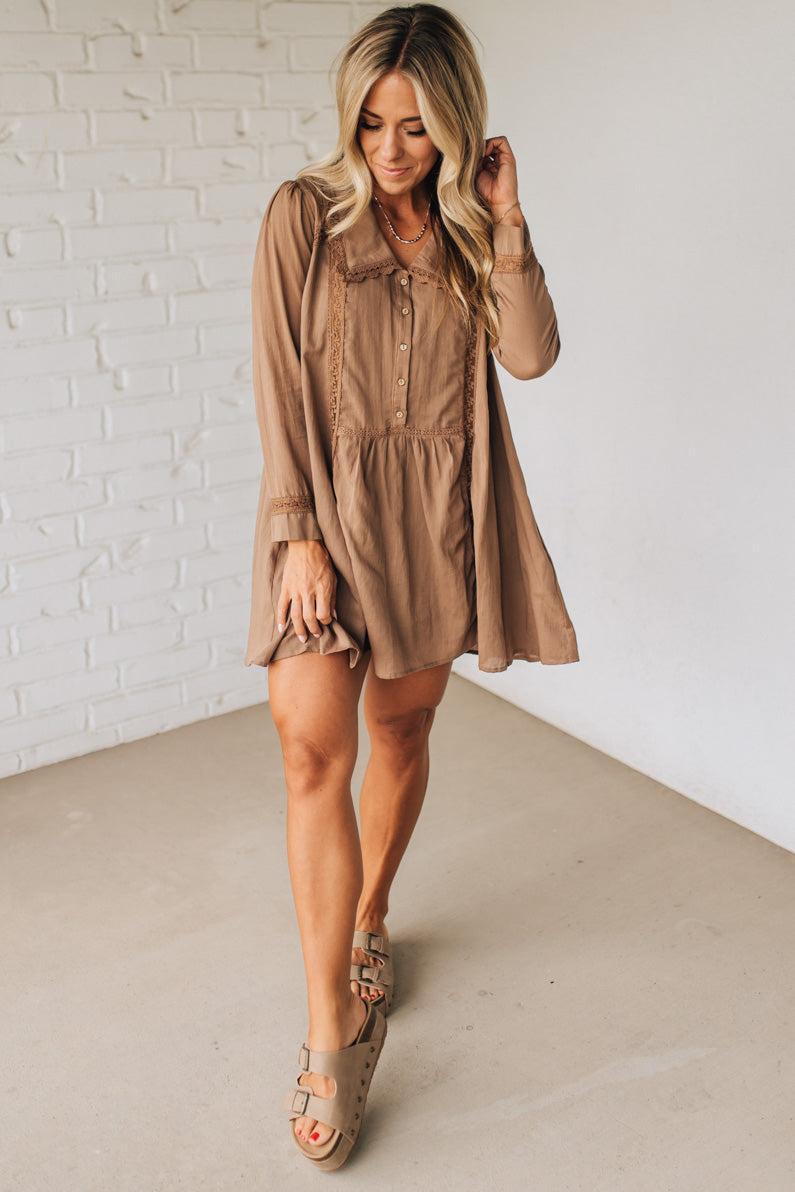Woman wearing a beige dress standing against a white brick wall.