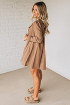 Woman wearing a beige dress standing against a white brick wall.