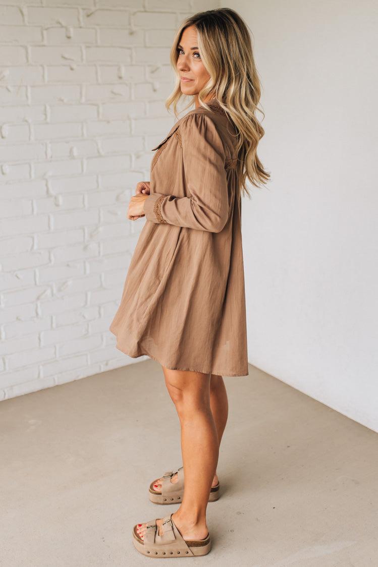 Woman wearing a beige dress standing against a white brick wall.