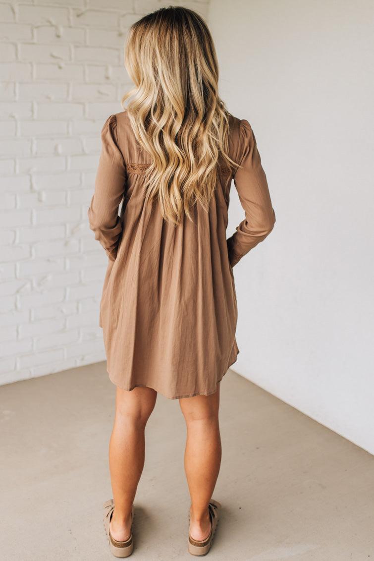 Woman wearing a brown dress standing against a white brick wall.