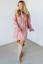 Woman wearing a pink denim dress and beige knee-high boots against a white brick wall.