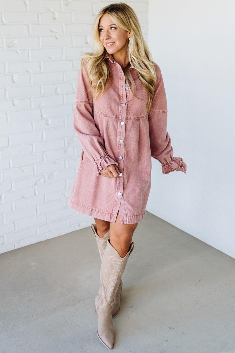 Woman wearing a pink denim dress and beige knee-high boots against a white brick wall.