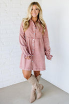 Woman wearing a pink denim dress with long sleeves against a white brick wall.