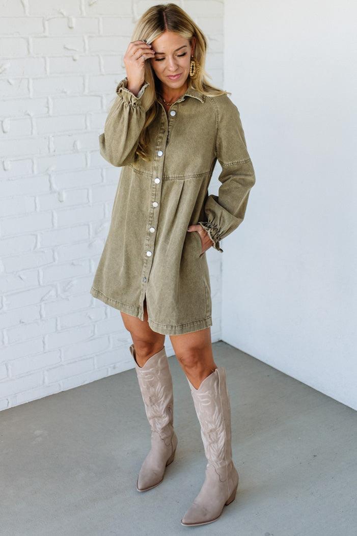 Woman wearing a green dress with long sleeves and buttons, paired with beige knee-high boots, against a white brick wall.