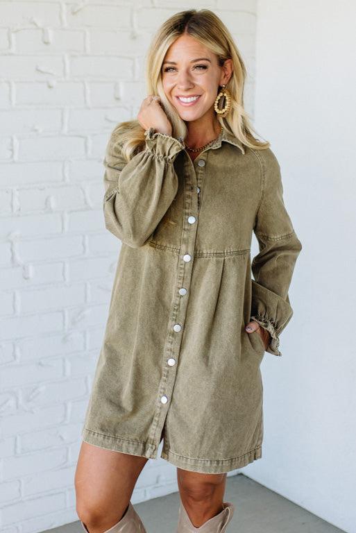 Woman wearing a green denim dress against a white brick wall.