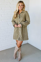 Woman wearing a green dress with long sleeves and beige knee-high boots against a white brick wall.