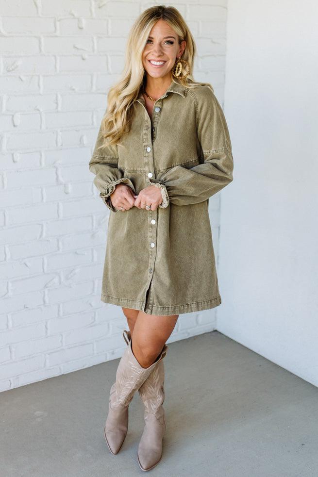 Woman wearing a green dress with long sleeves and beige knee-high boots against a white brick wall.