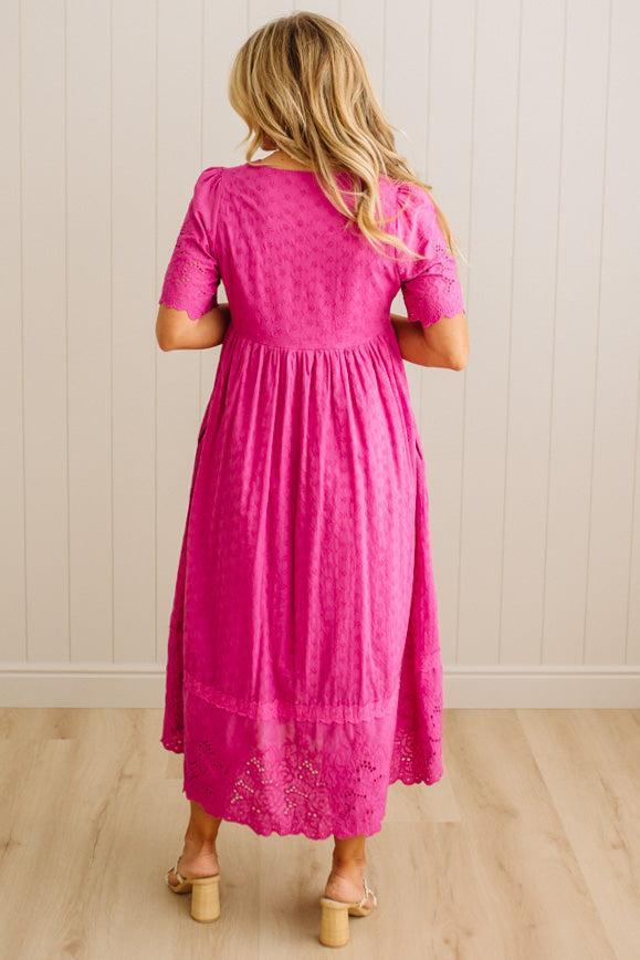 Woman wearing a pink dress with lace details, standing against a light wooden paneled wall.