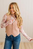 Woman wearing a pink ribbed vest over a floral blouse with blue jeans, standing against a neutral background.