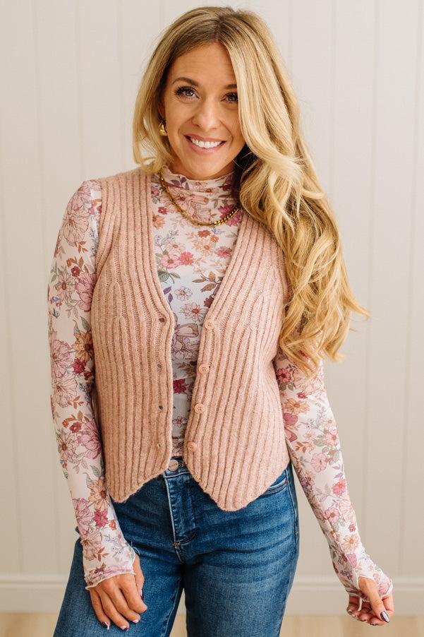 Woman wearing a pink ribbed vest over a floral long-sleeve shirt with blue jeans.