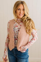 Woman wearing a floral long-sleeve top, pink ribbed vest, and blue jeans against a neutral background