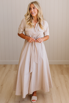 Woman wearing a beige dress standing in a room with wooden flooring and a light-colored wall.
