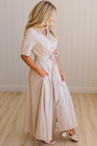 Woman wearing a long beige dress with a belt in a room with wooden flooring and a white wall.