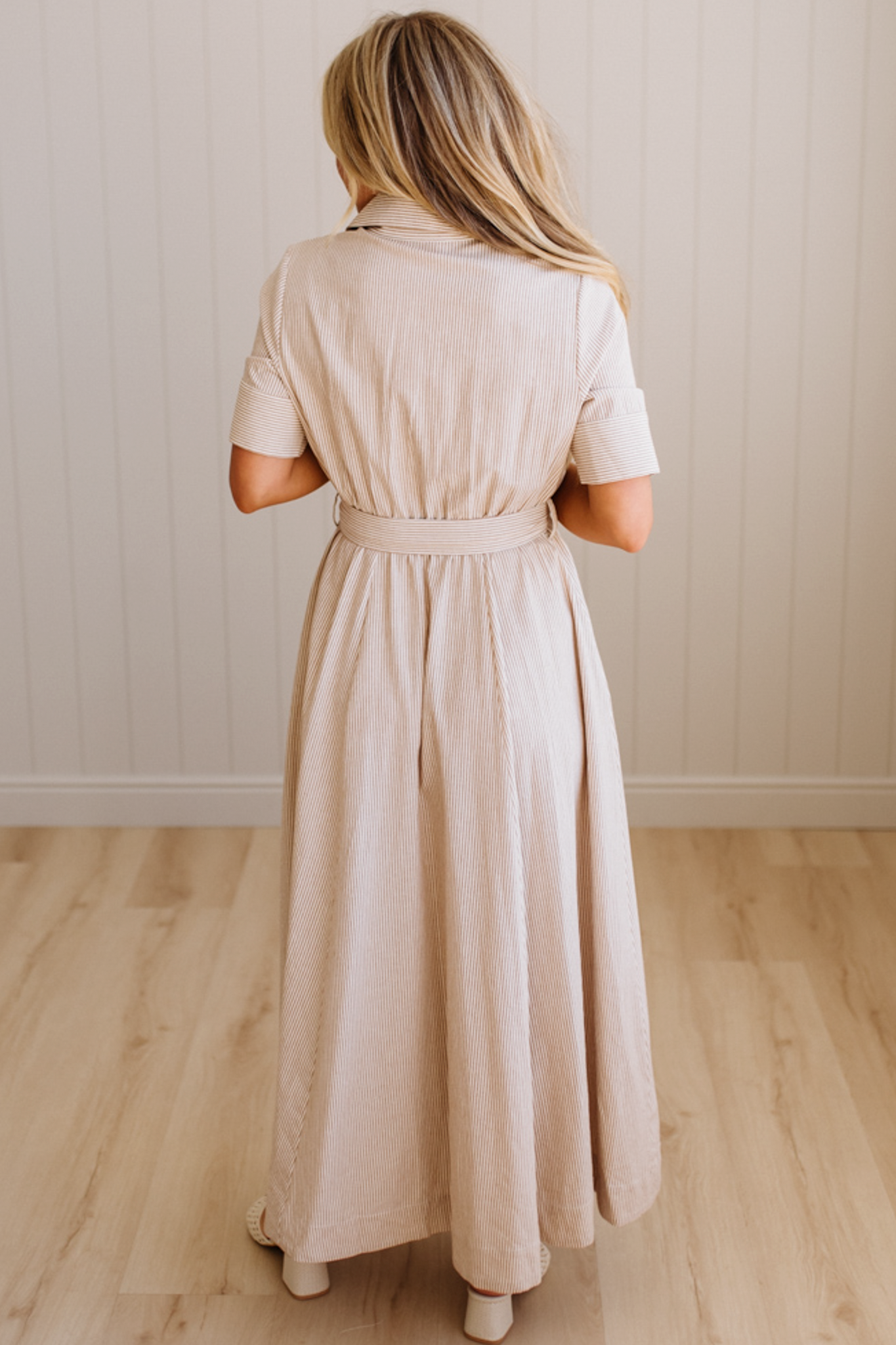 Woman wearing a beige dress standing in a room with wooden flooring and a white paneled wall.