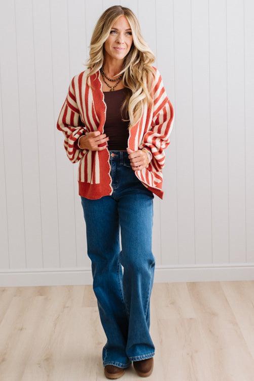 Blonde woman standing in a light studio wearing dark jeans and brown boots, with a rust orange and cream striped cardigan that has vertical lines and scallop edge trim.