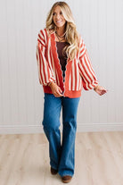 Blonde woman standing in a light studio wearing dark jeans and brown boots, with a rust orange and cream striped cardigan that has vertical lines and scallop edge trim.