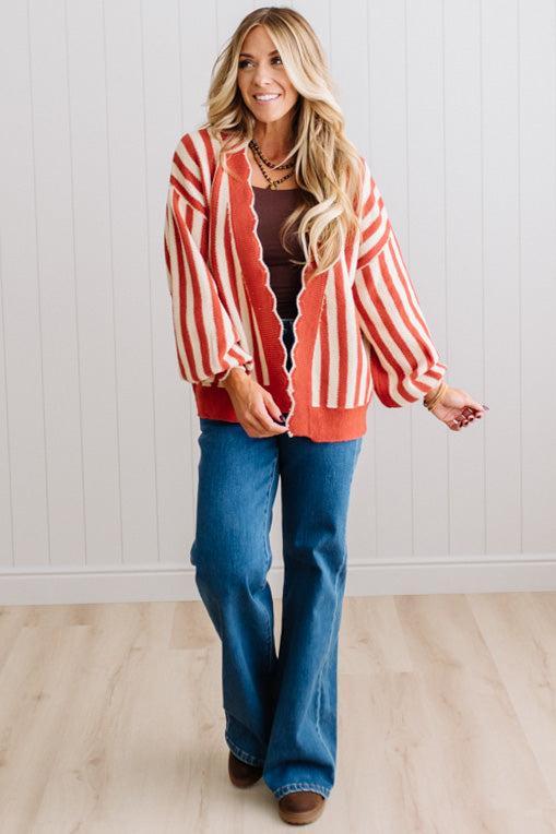 Blonde woman standing in a light studio wearing dark jeans and brown boots, with a rust orange and cream striped cardigan that has vertical lines and scallop edge trim.