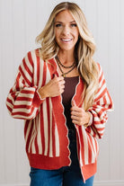 Blonde woman standing in a light studio wearing dark jeans and brown boots, with a rust orange and cream striped cardigan that has vertical lines and scallop edge trim.