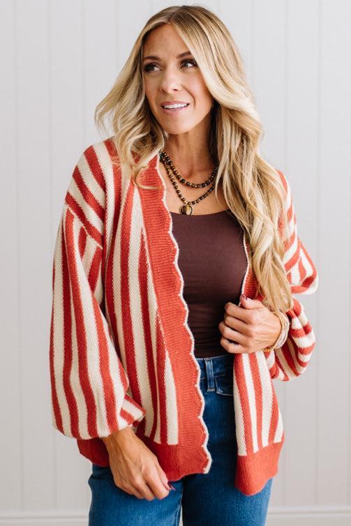 Blonde woman standing in a light studio wearing dark jeans and brown boots, with a rust orange and cream striped cardigan that has vertical lines and scallop edge trim.