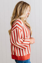 Blonde woman standing in a light studio wearing dark jeans and brown boots, with a rust orange and cream striped cardigan that has vertical lines and scallop edge trim.