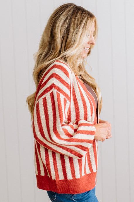 Blonde woman standing in a light studio wearing dark jeans and brown boots, with a rust orange and cream striped cardigan that has vertical lines and scallop edge trim.