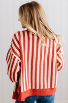 Blonde woman standing in a light studio wearing dark jeans and brown boots, with a rust orange and cream striped cardigan that has vertical lines and scallop edge trim.