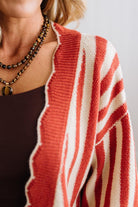 Blonde woman standing in a light studio wearing dark jeans and brown boots, with a rust orange and cream striped cardigan that has vertical lines and scallop edge trim.