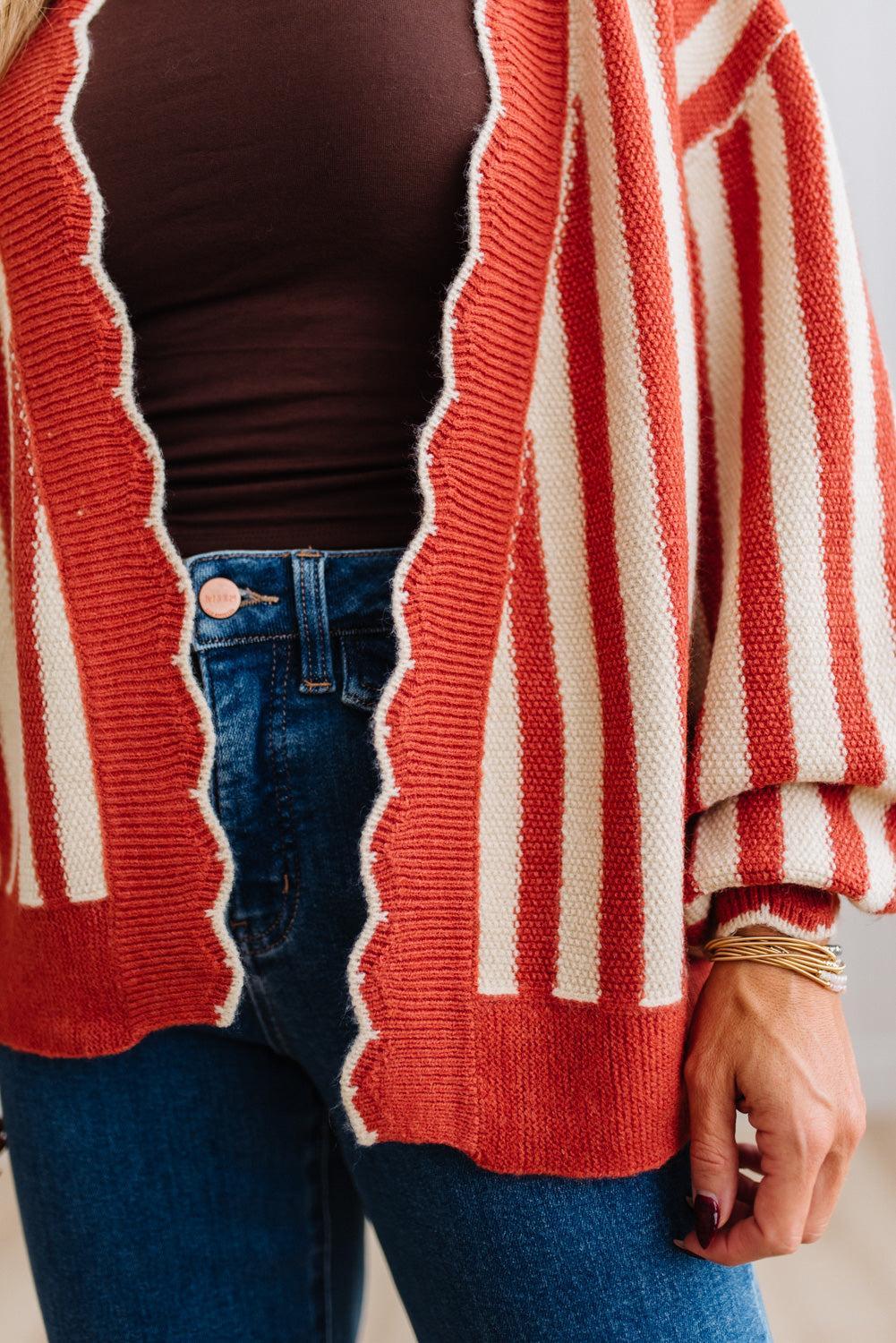 Blonde woman standing in a light studio wearing dark jeans and brown boots, with a rust orange and cream striped cardigan that has vertical lines and scallop edge trim.