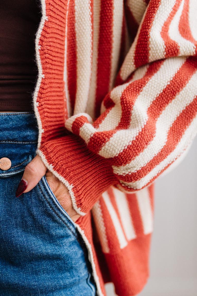 Blonde woman standing in a light studio wearing dark jeans and brown boots, with a rust orange and cream striped cardigan that has vertical lines and scallop edge trim.