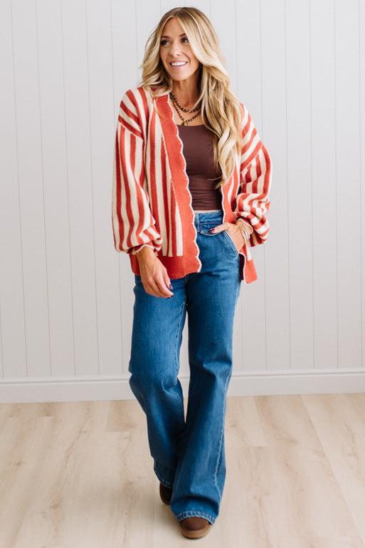 Blonde woman standing in a light studio wearing dark jeans and brown boots, with a rust orange and cream striped cardigan that has vertical lines and scallop edge trim.