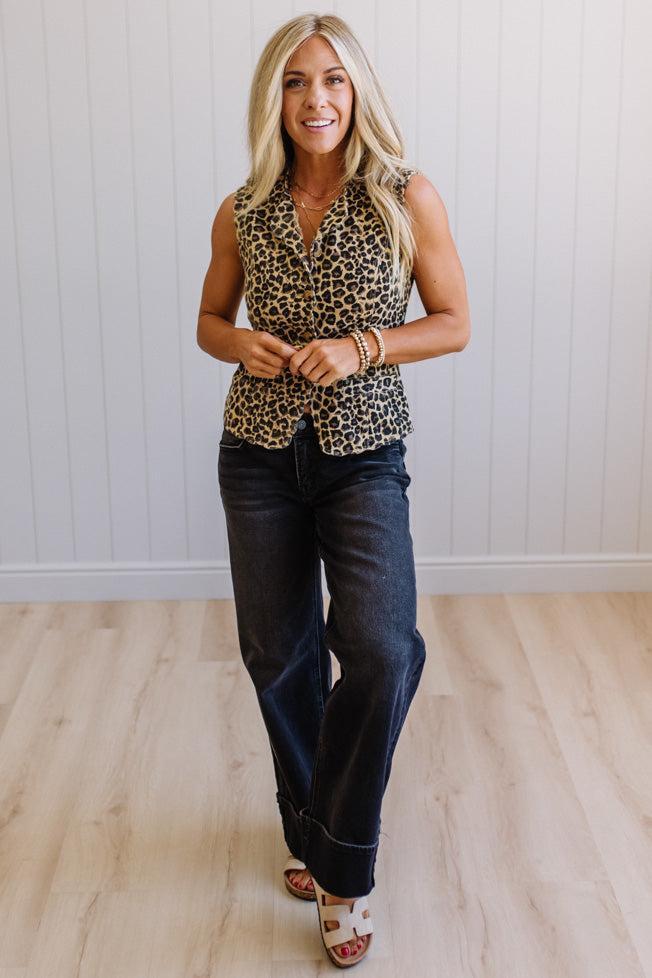 Woman wearing a leopard print top and blue jeans standing against a white paneled wall.