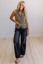 Woman wearing a leopard print sleeveless top and blue jeans standing against a white wall.