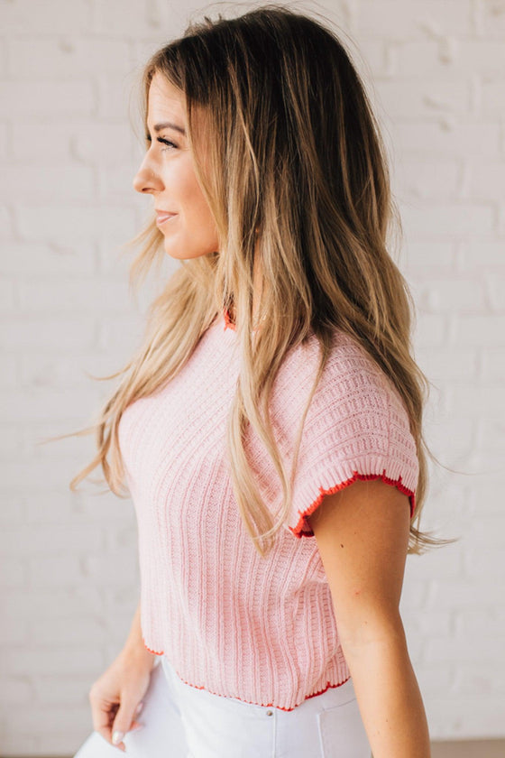 Blonde woman wearing a textured pink sweater with scalloped red trim.