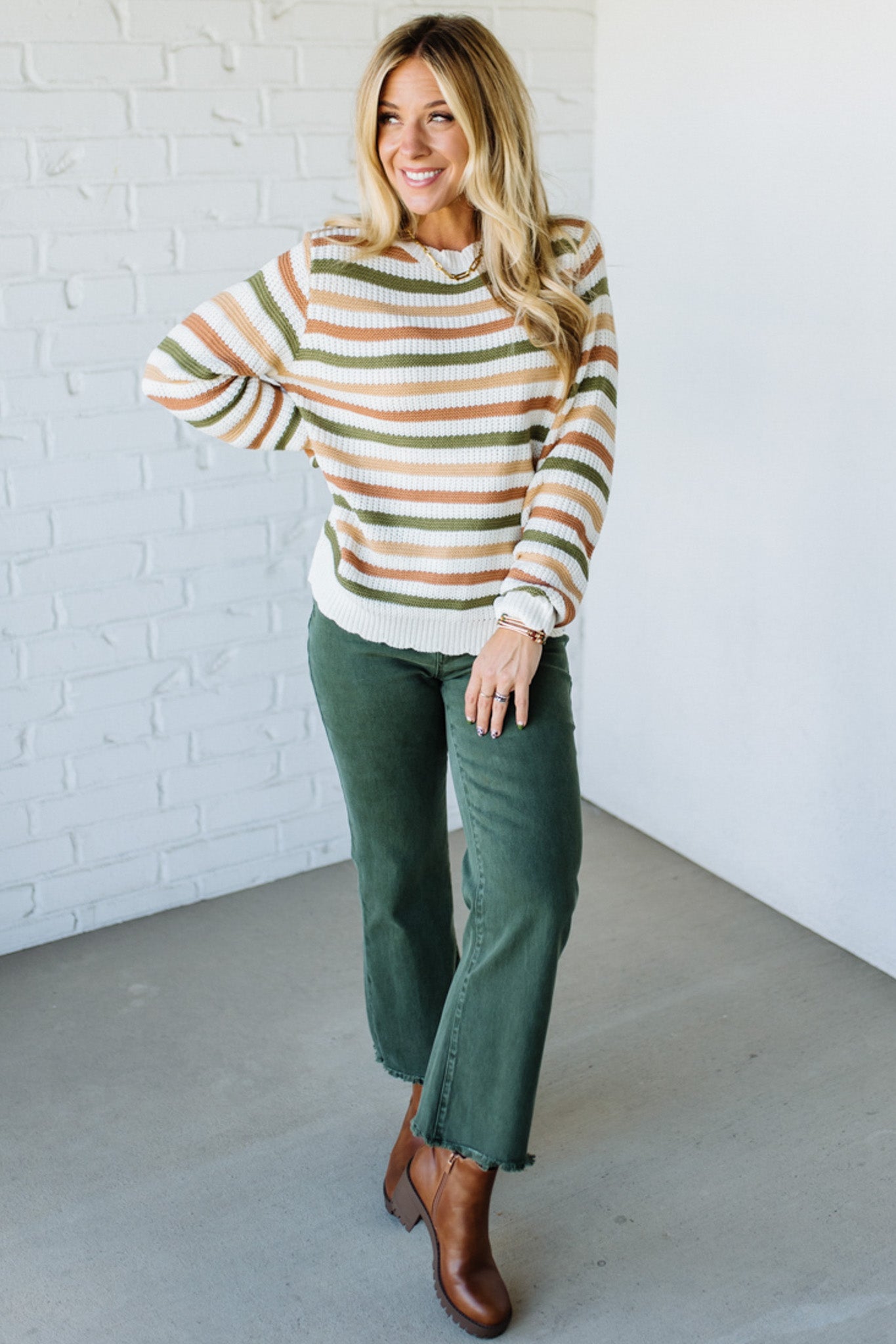 Woman wearing a striped sweater and green pants against a white brick wall.