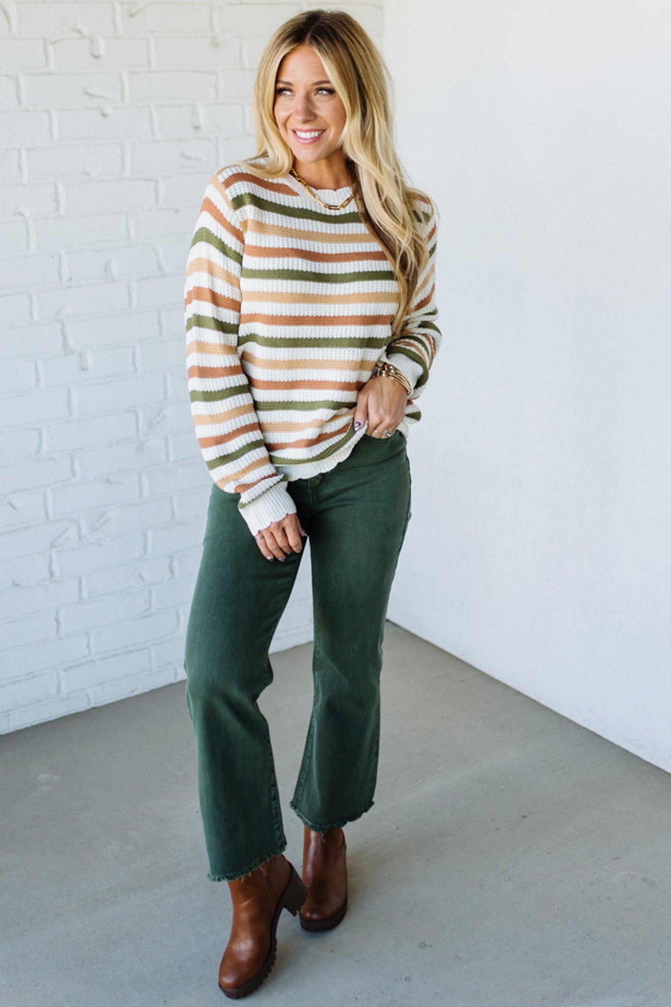 Woman wearing a striped sweater and green pants against a white brick wall.