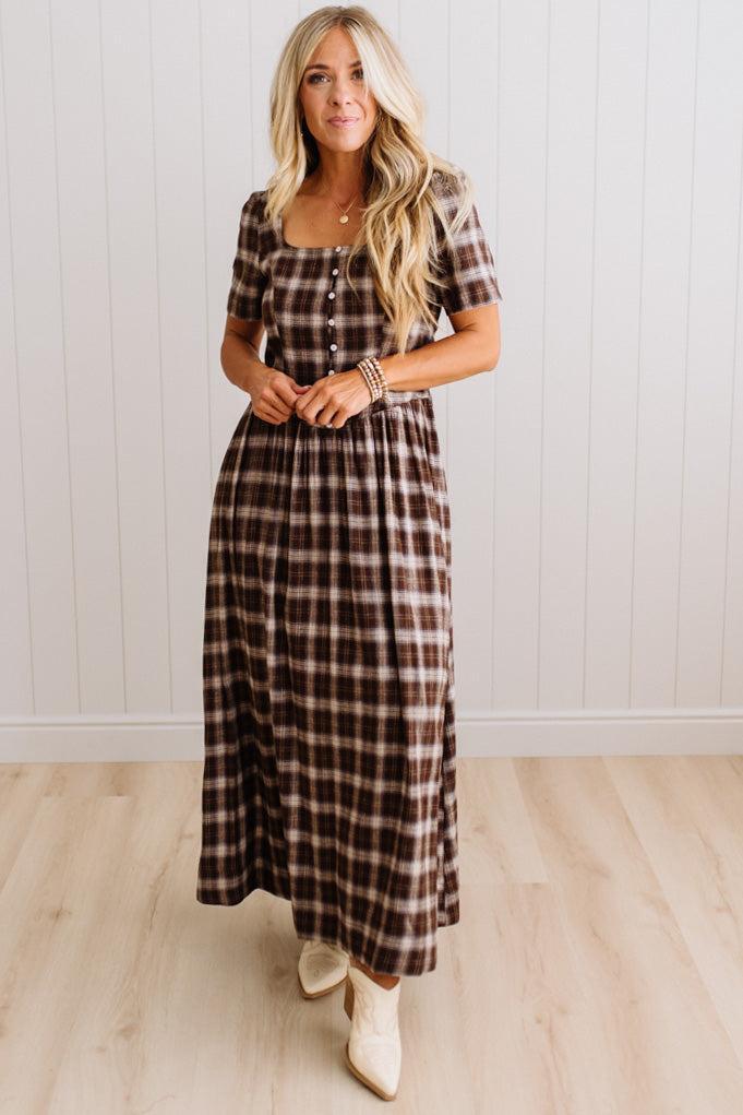 A woman modeling a brown and beige plaid short-sleeve maxi dress with a button-up front, standing in front of a white paneled wall and light wood floor.