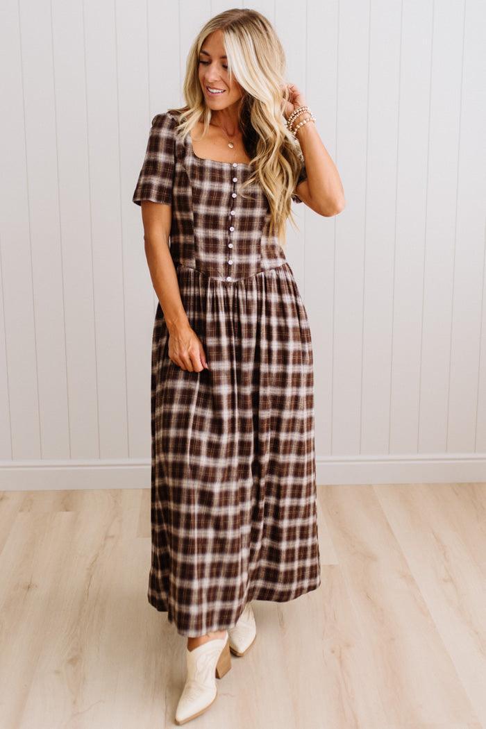A woman modeling a brown and beige plaid short-sleeve maxi dress with a button-up front, standing in front of a white paneled wall and light wood floor.