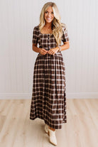 A woman modeling a brown and beige plaid midi dress with short sleeves, a square neckline, and a button-up front, standing in front of a white paneled wall and light wood floor.