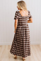A woman wearing a brown and beige plaid maxi dress with puff sleeves and a square neckline, shown from the back in a bright indoor setting with white paneled walls and light wood flooring.
