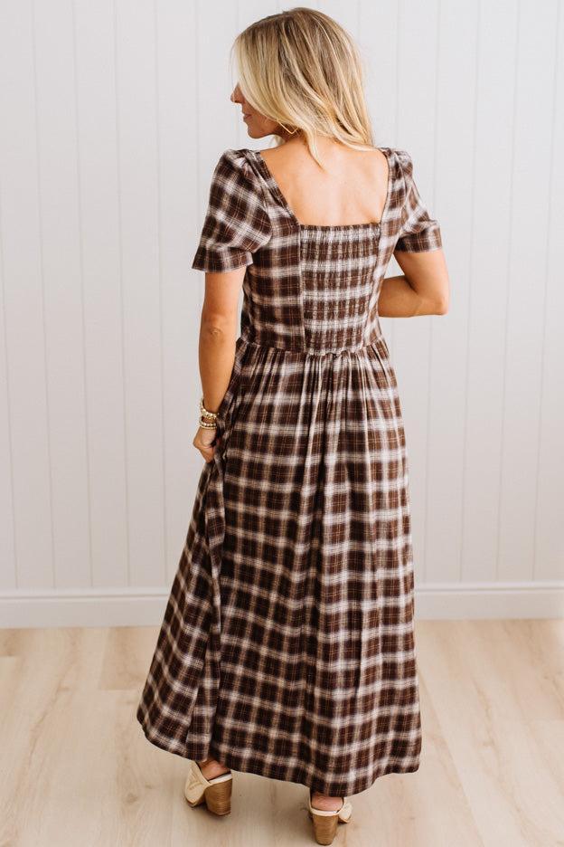 A woman wearing a brown and beige plaid maxi dress with puff sleeves and a square neckline, shown from the back in a bright indoor setting with white paneled walls and light wood flooring.
