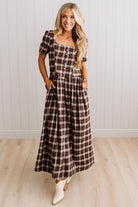 A woman modeling a brown and beige plaid maxi dress with short puff sleeves, a square neckline with button details, and side pockets, standing in front of a white paneled wall and light wood floor.
